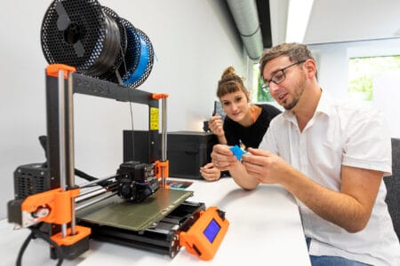 Eine junge Frau und ein junger Mann mit Brille sitzen an einem 3D-Drucker.