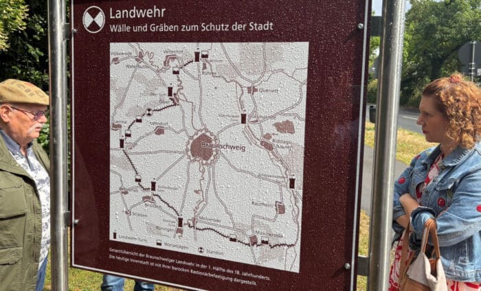 Die Frontseite des neuen BLIK-Schildes am Lammer Holz zeigt den Verlauf des Landwehrs. Links im Bild Rudolf Rischmann von der Schwabe-Stiftung Kuchelberg, rechts Sabrina Brandes, Ortsheimatpflegerin Lehndorf. Foto: Stadtheimatpflege